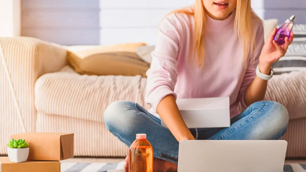 Excited young woman is doing online shopping at home. She is using laptop and holding perfume